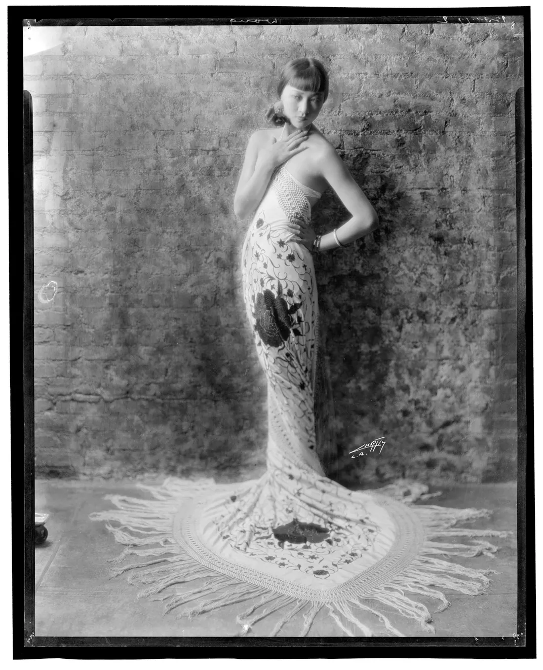 Black-and-white portrait of Anna May Wong standing against a wall, wearing an elaborate dress decorated in a flower moti