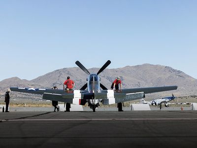 Speedball Alice, a restored dark green P-51D airplane with white stripes, is parked on a tarmac in the Reno desert, with mountains and blue sky in the background.