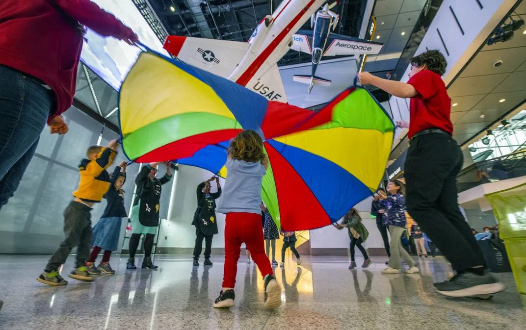A group of children play with a parachute in a museum with airplanes overhead
